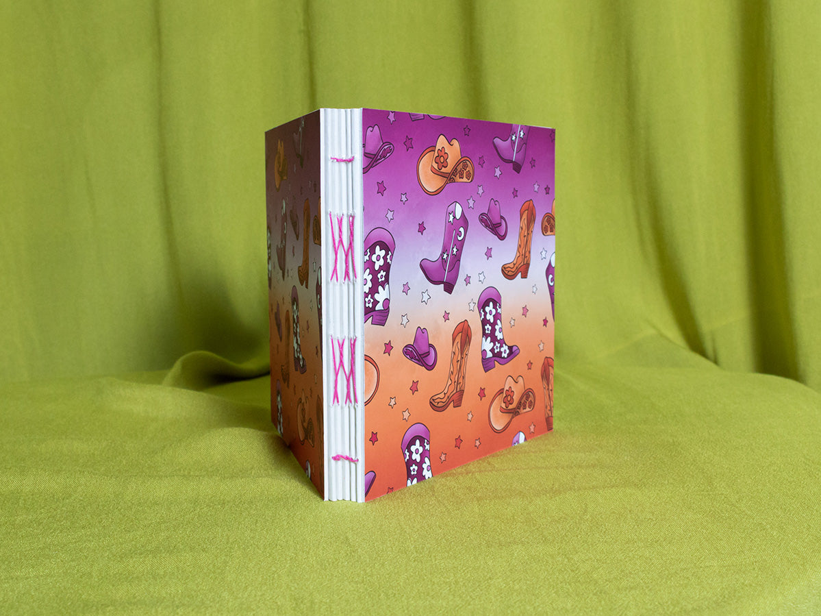 Open spine square sketchbook with colourful cowgirl design on a green fabric background