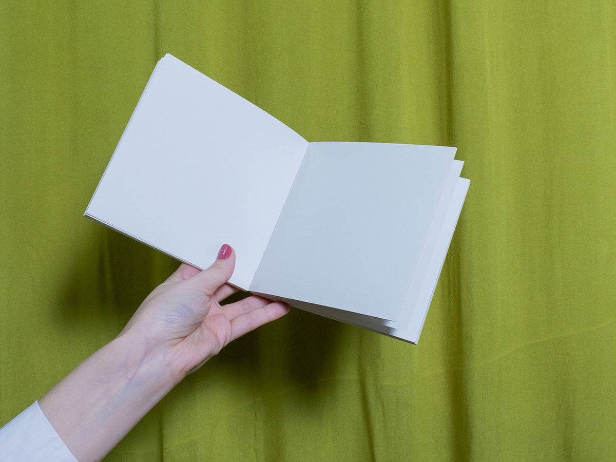 Hand holding a blank square sketchbook against a green curtain background