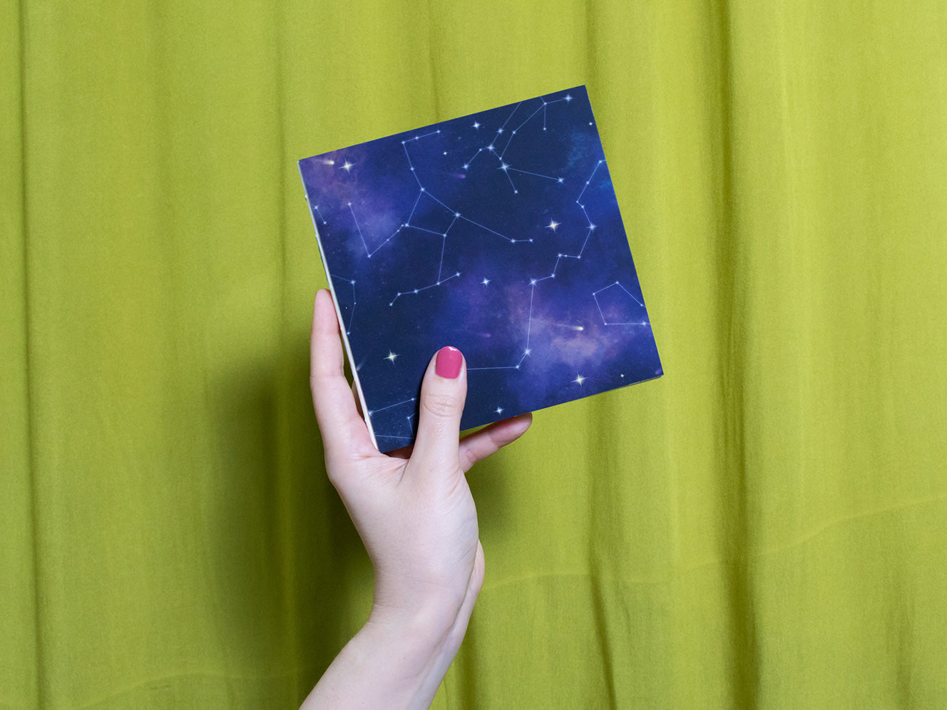 Hand holding a square sketchbook with star constellations against a green curtain background