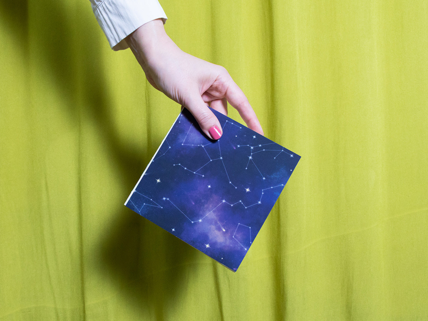 Hand holding a square sketchbook with star constellations against a yellow curtain background