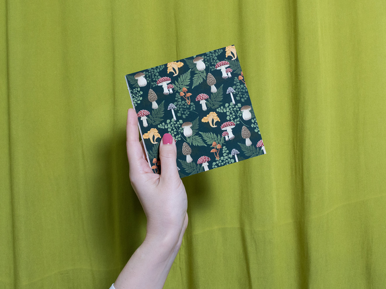 Hand holding a square sketchbook with a mushroom pattern against a green curtain background