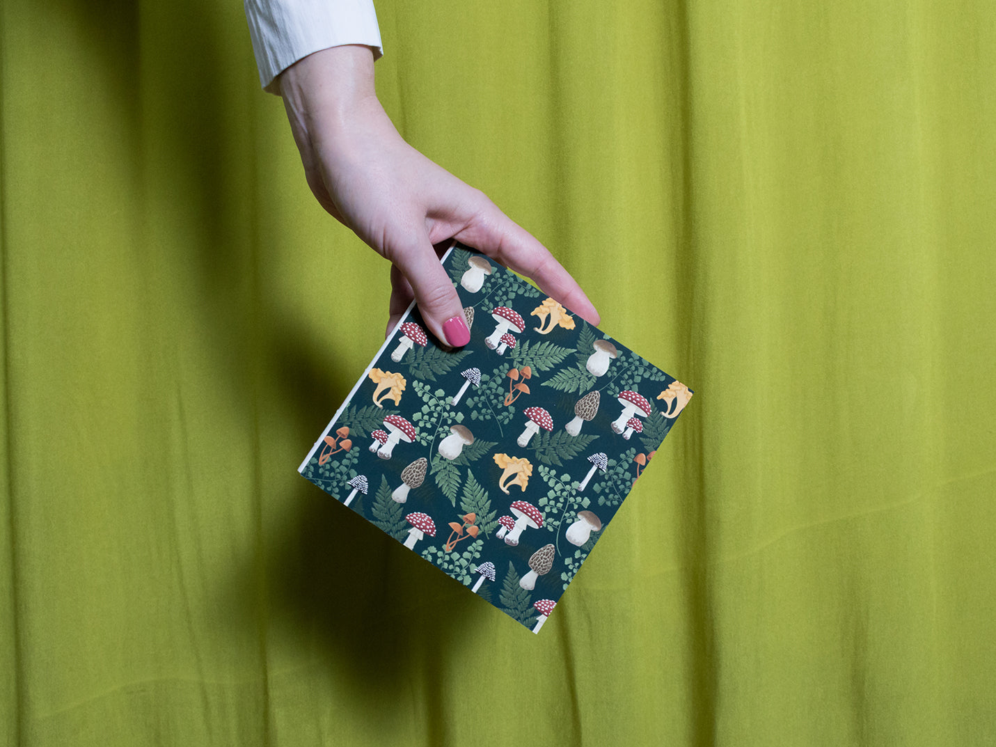 Hand holding a square sketchbook with a mushroom cover against a green curtain background